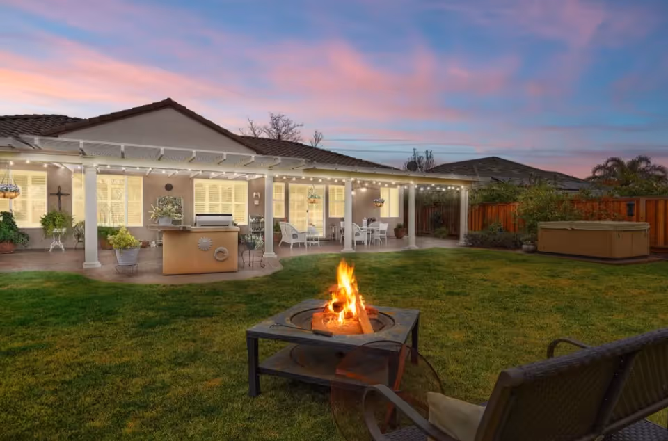 A backyard patio area at dusk with a fire pit burning in the foreground, a bench with a cushion nearby, and a covered patio attached to a house with outdoor furniture, hanging plants, and string lights. The sky is colorful with shades of pink and blue.