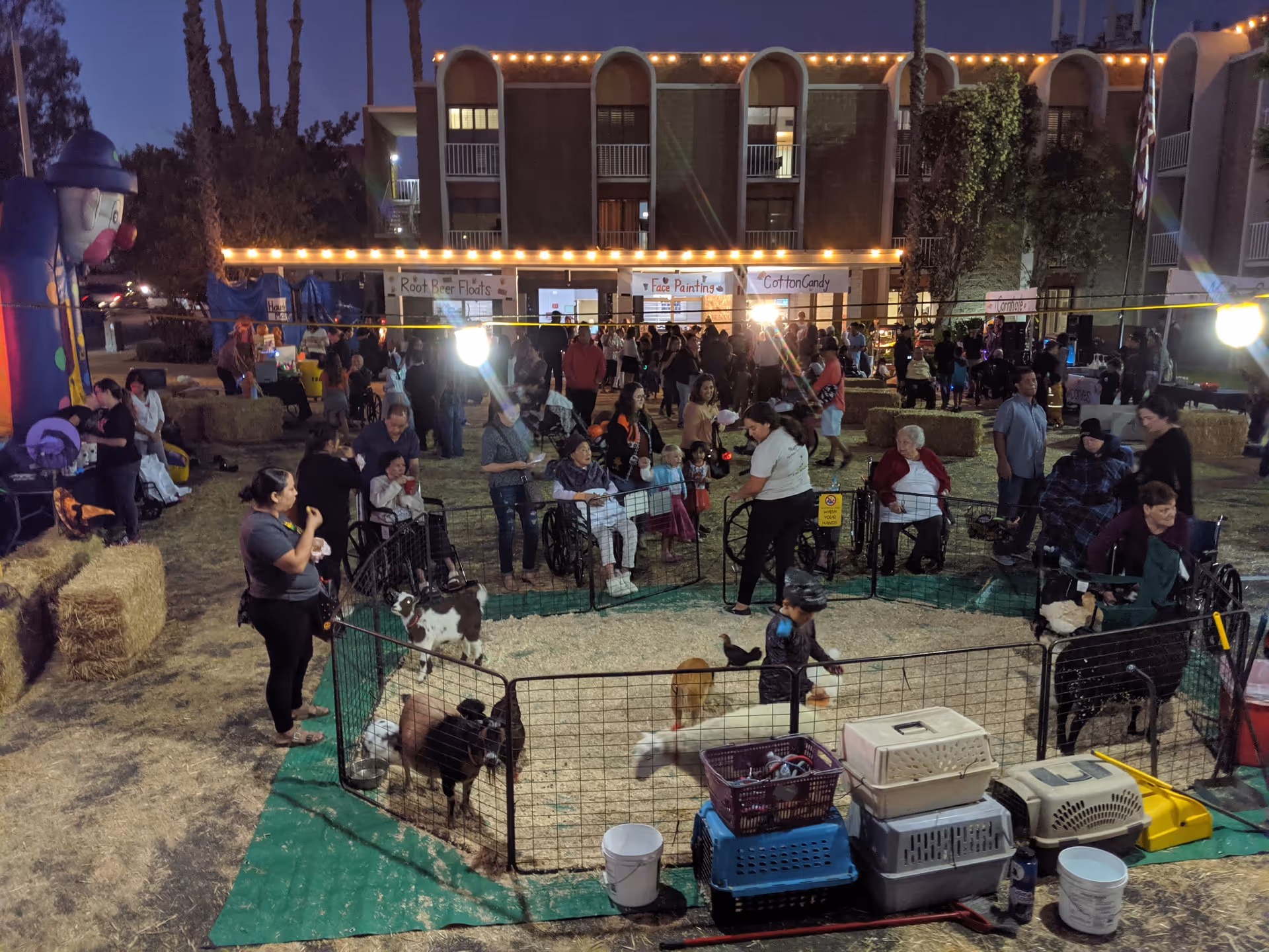 An outdoor evening event at The Pavilion at Sunny Hills with people of various ages gathered around a small petting zoo enclosure containing goats and chickens. There are hay bales arranged around the area, a large inflatable clown on the left, and several booths in the background with signs for Root Beer Floats, Face Painting, and Cotton Candy. The building behind is decorated with string lights.