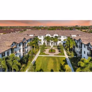 Aerial view of a large senior living facility with a central courtyard featuring a circular garden and palm trees, surrounded by multi-story buildings with tiled roofs under a sunset sky.