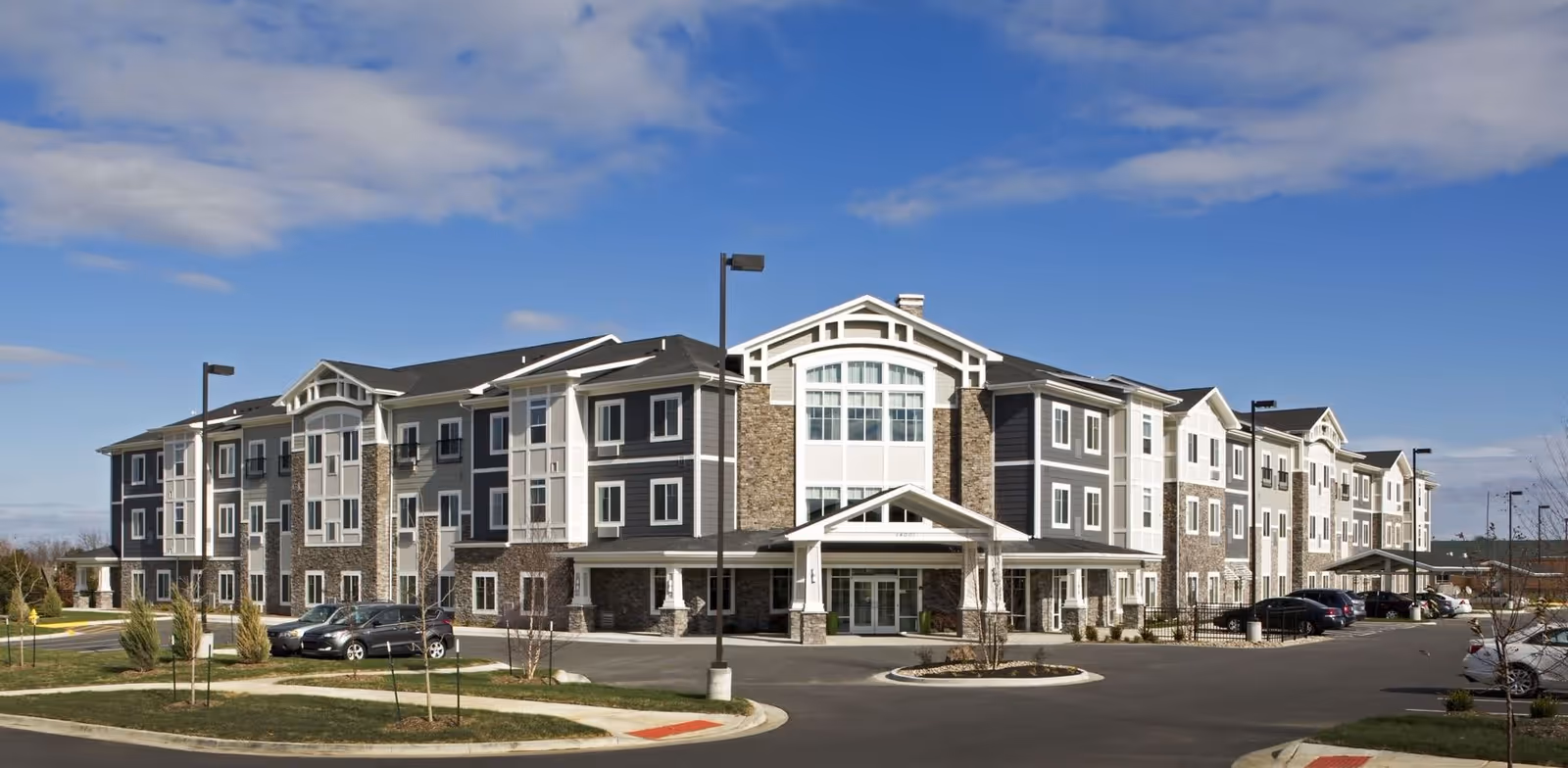 Exterior view of a large, modern three-story senior living facility building with a mix of stone and siding facade, multiple windows, and a covered entrance. Several cars are parked in the parking lot in front of the building under a partly cloudy blue sky.