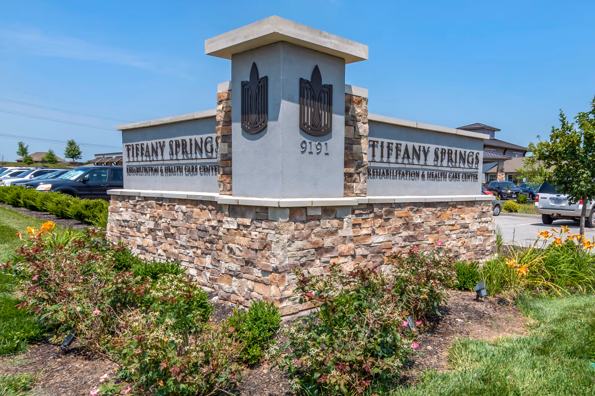 Stone and concrete sign at the entrance of Tiffany Springs Rehabilitation & Health Care Center with landscaping and parked cars in the background under a clear blue sky.