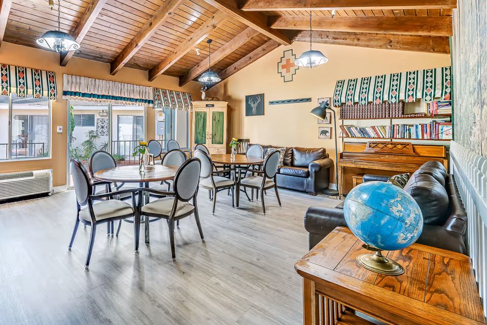 A bright and spacious common area with wooden vaulted ceilings and large windows. The room features round tables with chairs, two leather sofas, a wooden piano with shelves of books above it, and a small wooden table with a globe on it. The windows have patterned valances, and there are decorative wall hangings including a deer head silhouette and a geometric design.