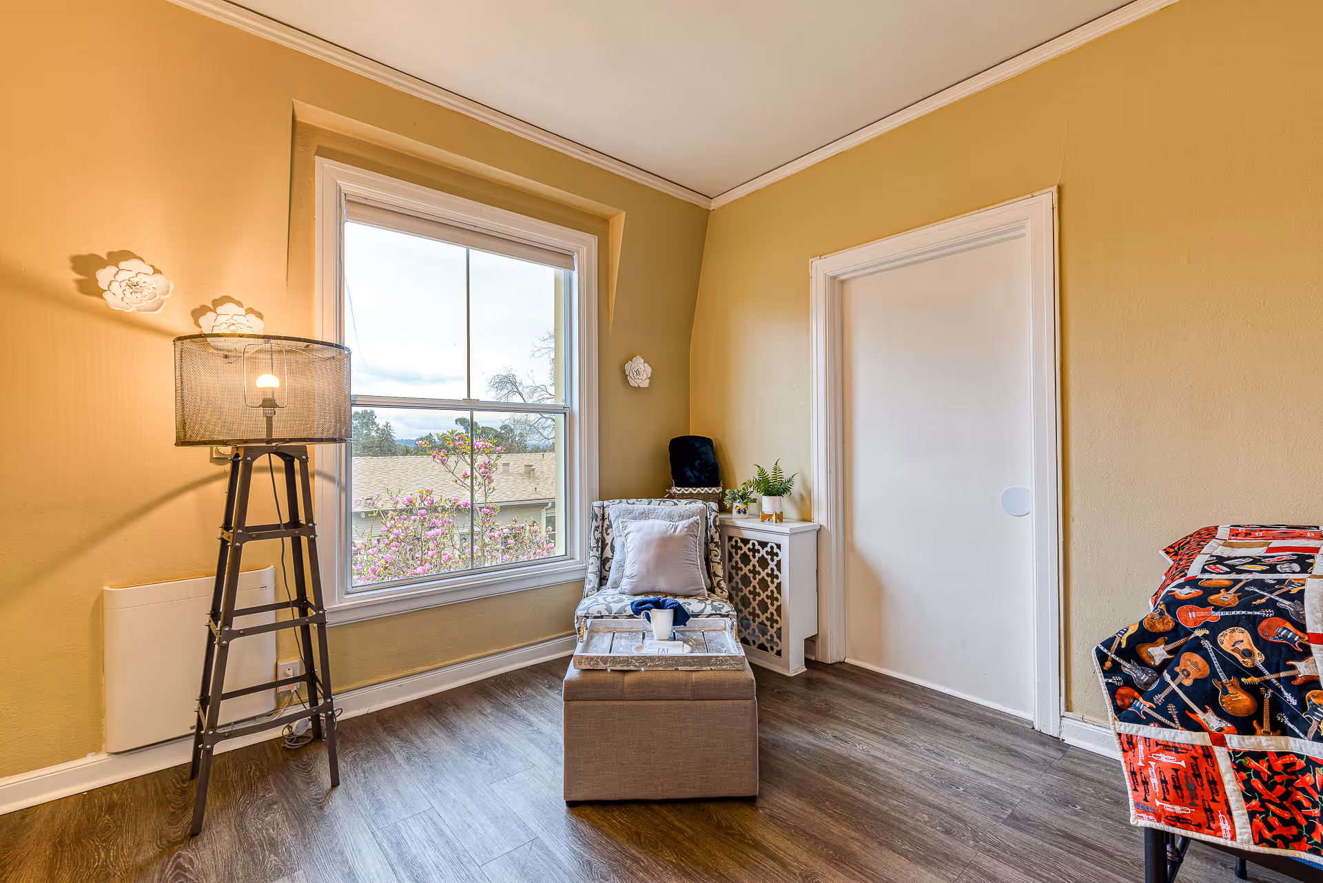 A cozy corner of a senior living bedroom with a patterned armchair, a small ottoman with a tray holding a cup, a floor lamp, a window showing blooming pink flowers outside, and part of a bed with a colorful quilt featuring guitars.