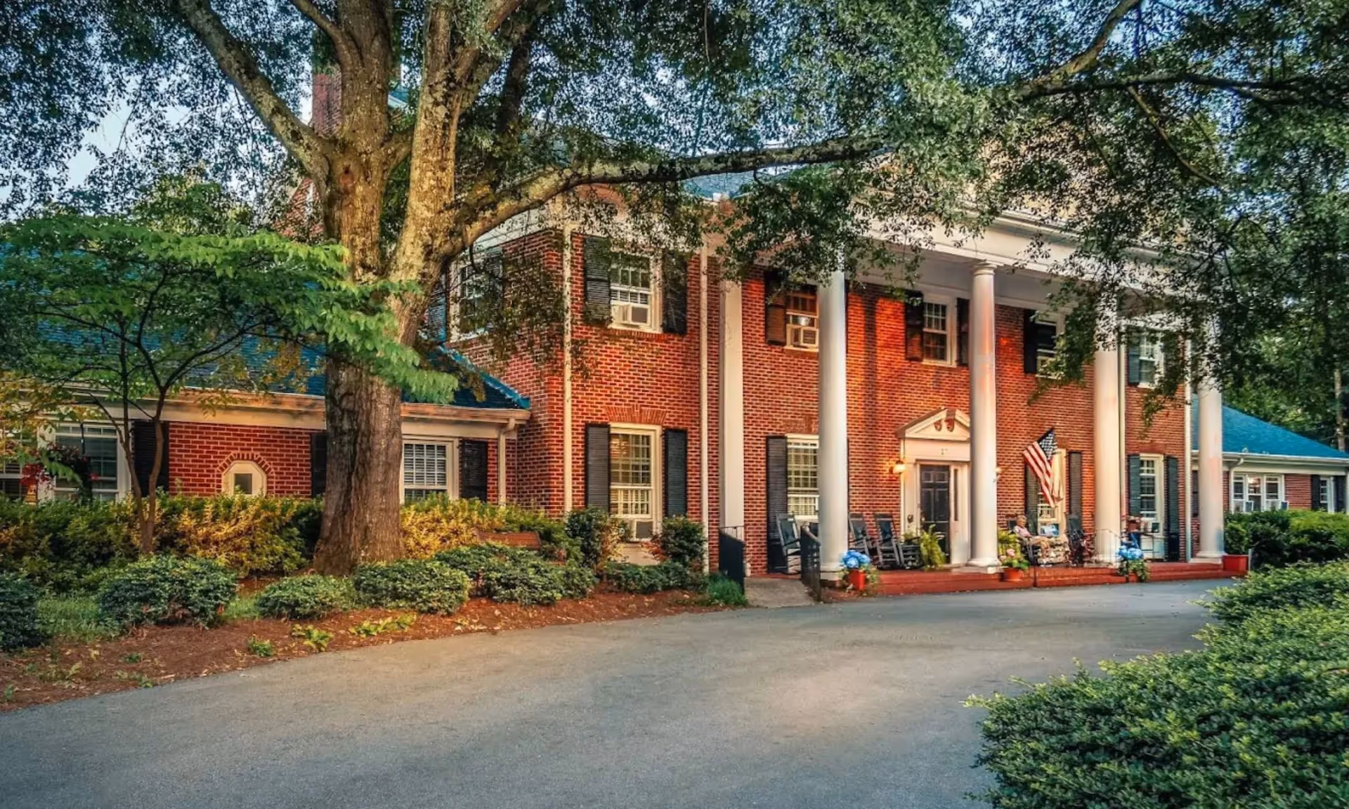 Front view of a brick retirement home with white columns, a flag, and landscaped surroundings.