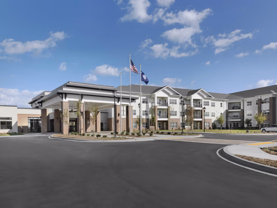 Exterior view of a large, modern senior living facility building with a covered entrance, three flagpoles with flags, and a circular driveway under a partly cloudy blue sky.