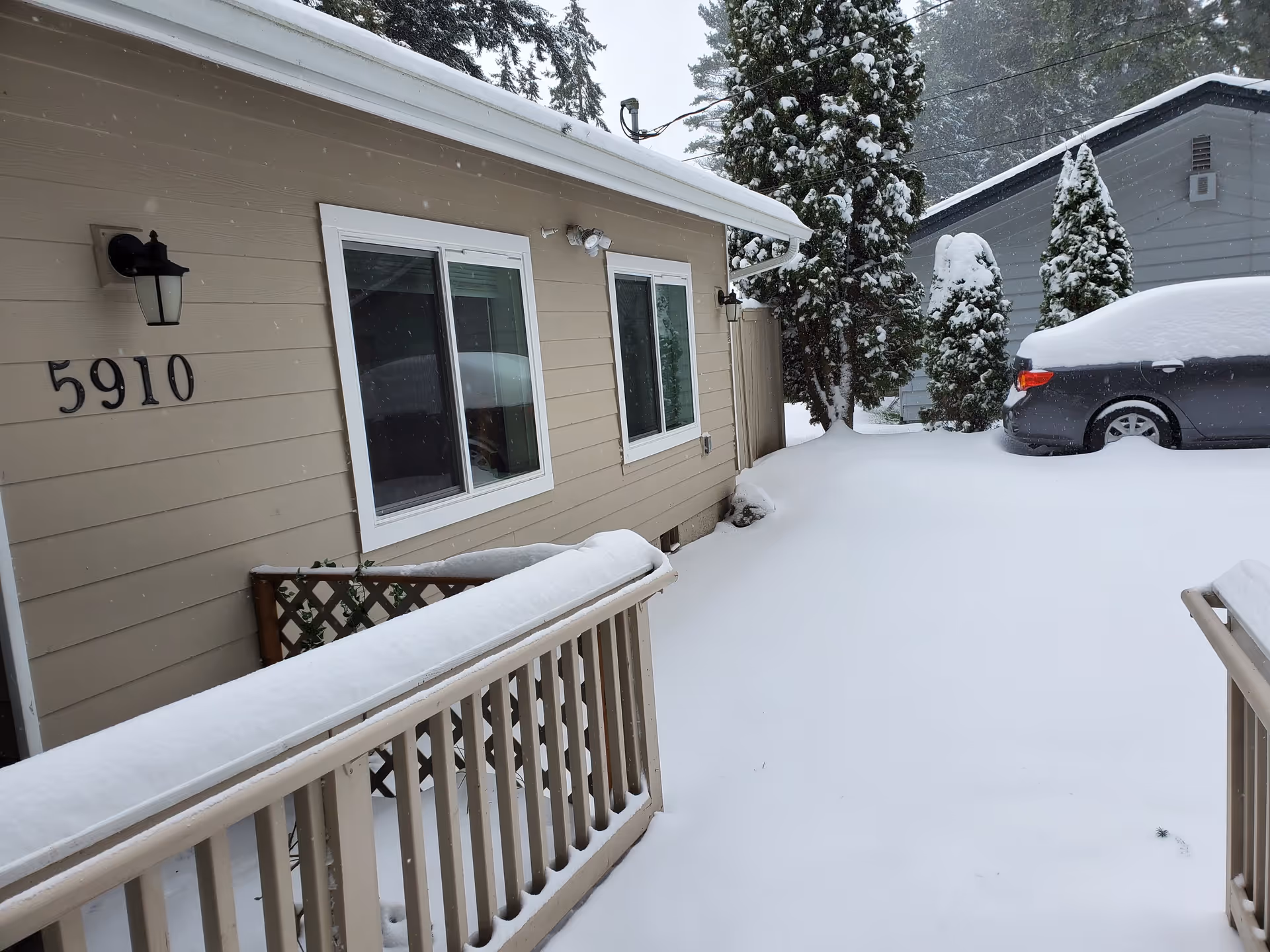Snow-covered outdoor area beside a beige building with the number 5910 on the wall. There is a wooden railing also covered in snow, a parked car covered in snow, and snow-covered trees in the background.