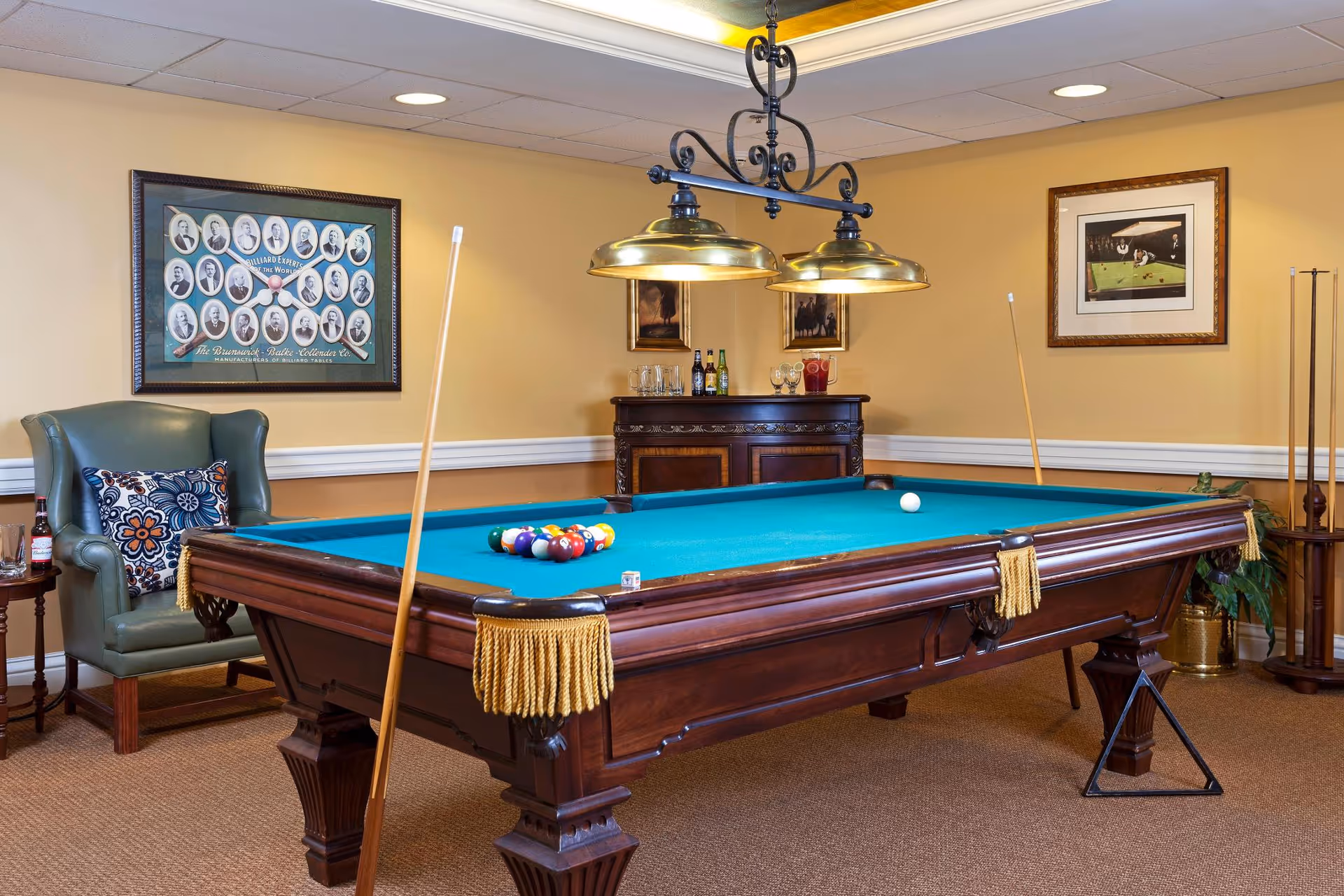 A well-appointed billiards room featuring a wooden pool table with teal felt, hanging brass lights, cue sticks, an armchair, and framed artwork on the walls.