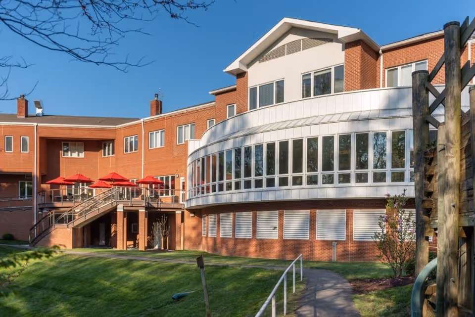 Exterior view of a multi-story brick building with large curved windows and a balcony area featuring red umbrellas. There is a grassy lawn and a paved walkway with a railing leading towards the building under a clear blue sky.