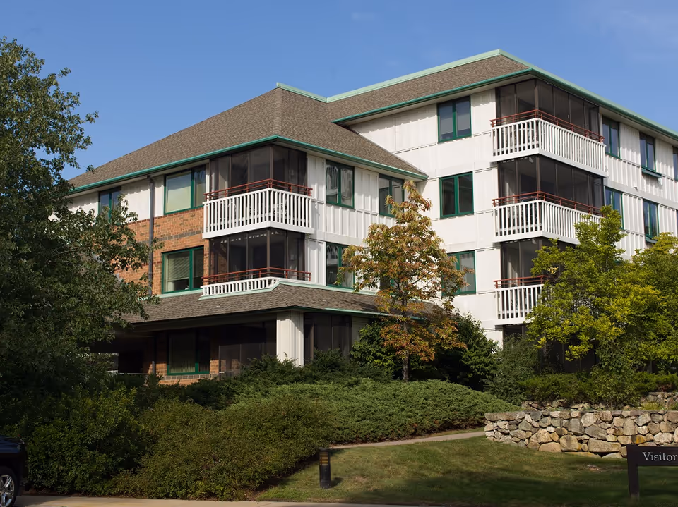 Exterior view of a multi-story senior living facility building with screened balconies, surrounded by trees and shrubs under a clear blue sky.