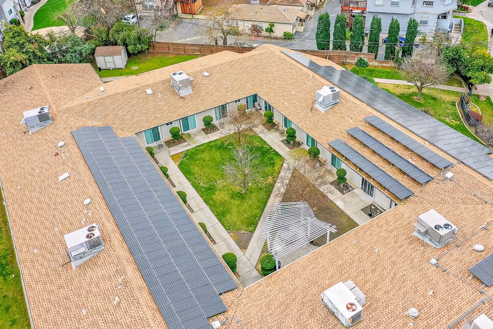 Aerial view of a single-story assisted living facility with a central courtyard featuring green grass, a tree, shrubs, and a white pergola. The building has a tan roof with multiple solar panels installed and several HVAC units on top. Surrounding the courtyard are walkways and entrances to the facility.
