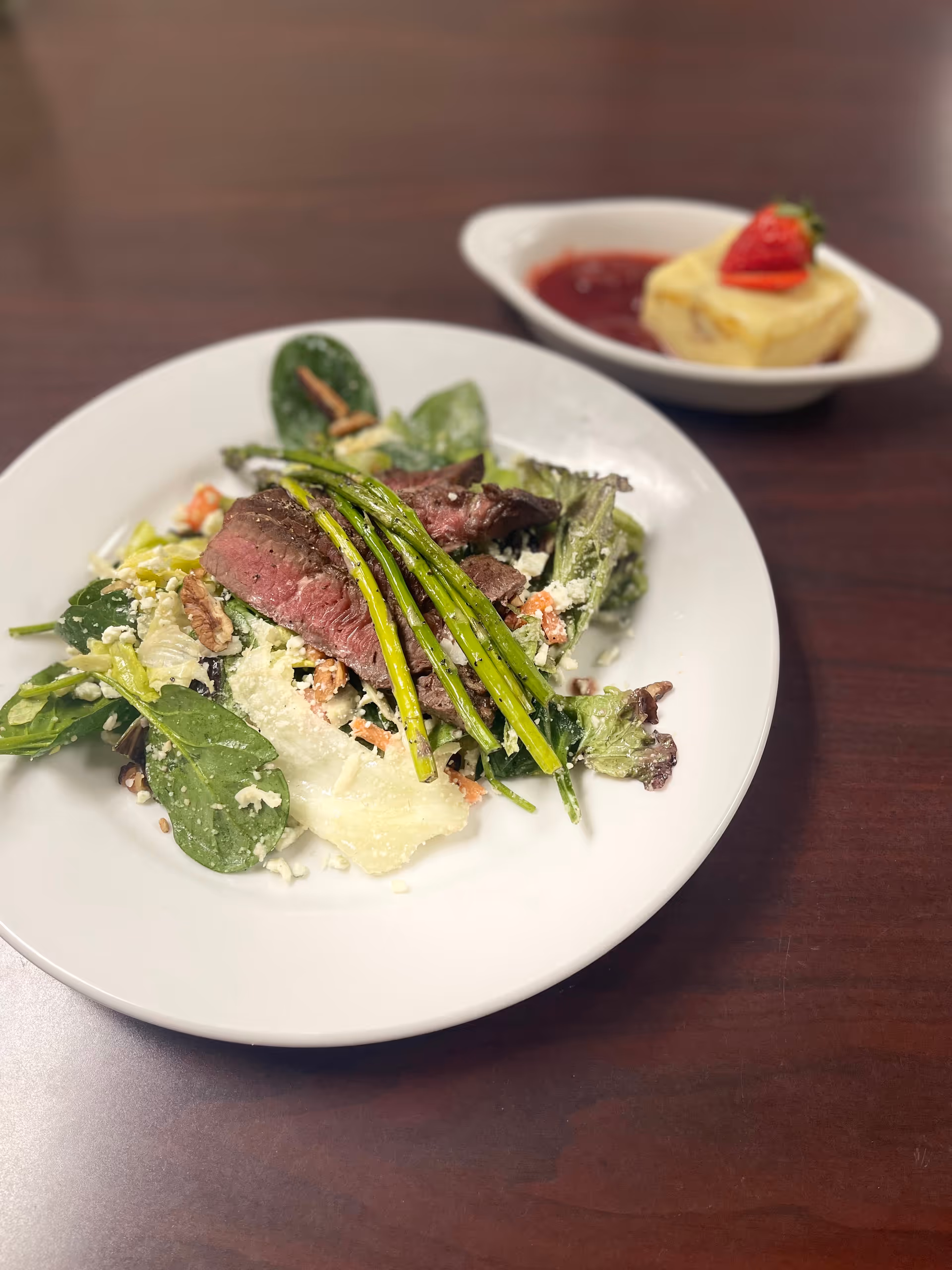 A plate of salad with mixed greens, slices of medium-rare steak, and grilled asparagus on top. In the background, there is a small dish with a dessert topped with a strawberry and a red sauce.