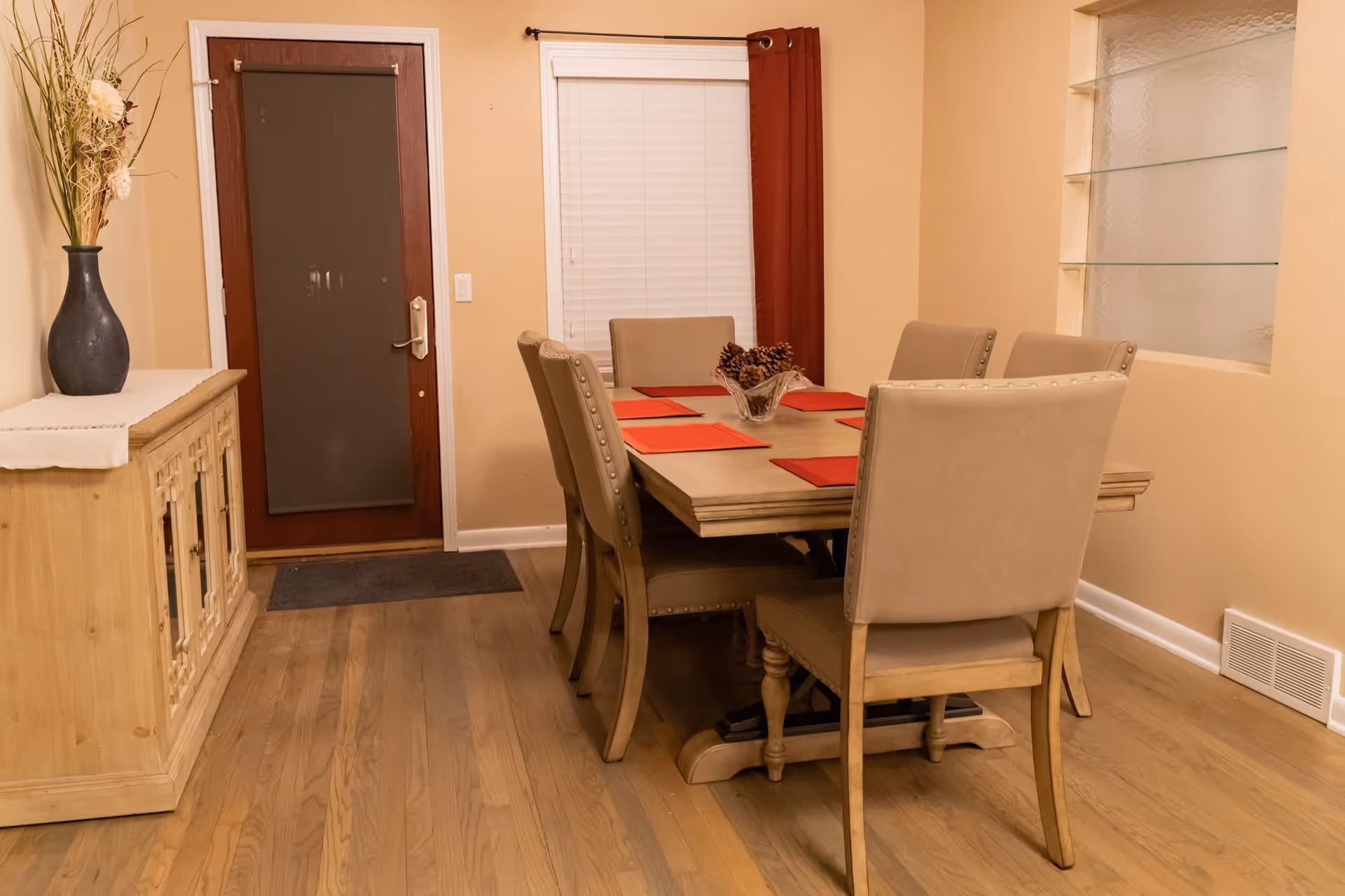 A dining room with a wooden table set with six beige upholstered chairs. The table has red placemats and a decorative centerpiece with pinecones. There is a wooden sideboard with a vase of dried flowers on the left, a door with a dark screen, a window with white blinds and red curtains, and a built-in glass shelf on the right wall.