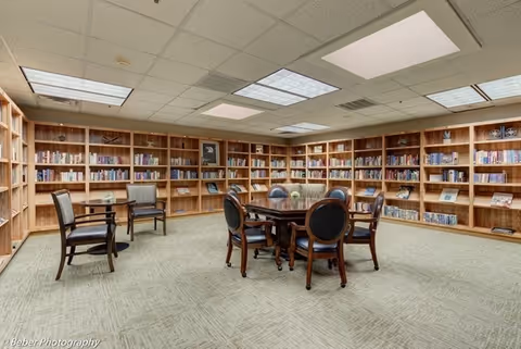 A spacious library room with wooden bookshelves lining the walls filled with books. In the center, there is a round wooden table surrounded by six chairs with dark cushions. Two additional chairs with armrests are placed near a small table on the left side. The room has a carpeted floor and a ceiling with fluorescent lighting panels.