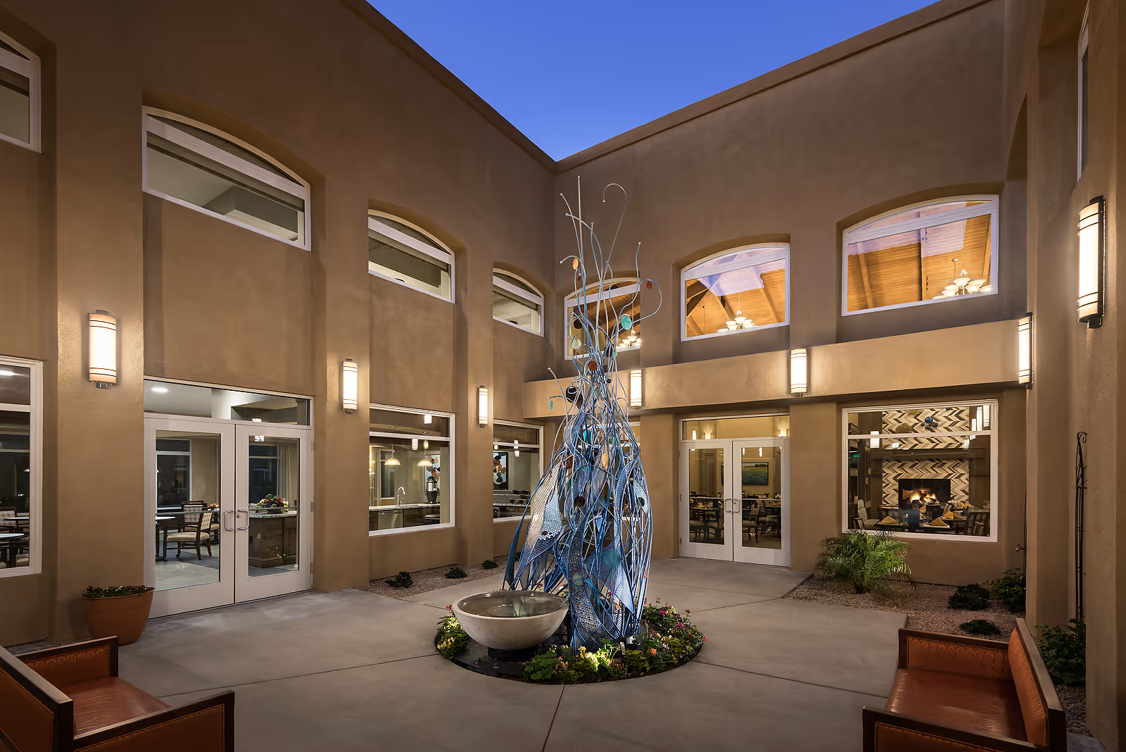 Enclosed courtyard at dusk featuring a central metal sculpture and fountain, seating, and surrounding windows and doors into the building.