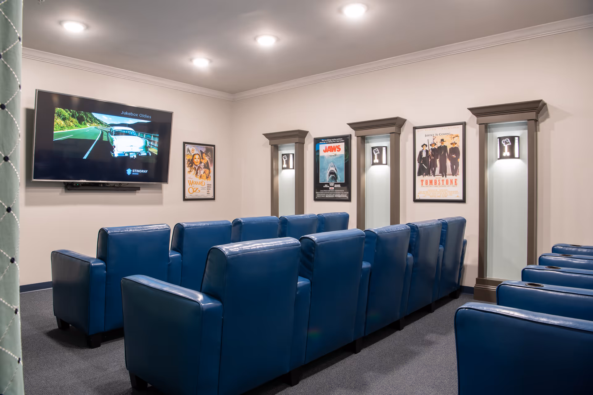 Small screening room with rows of blue recliner seats facing a wall-mounted TV and framed movie posters.