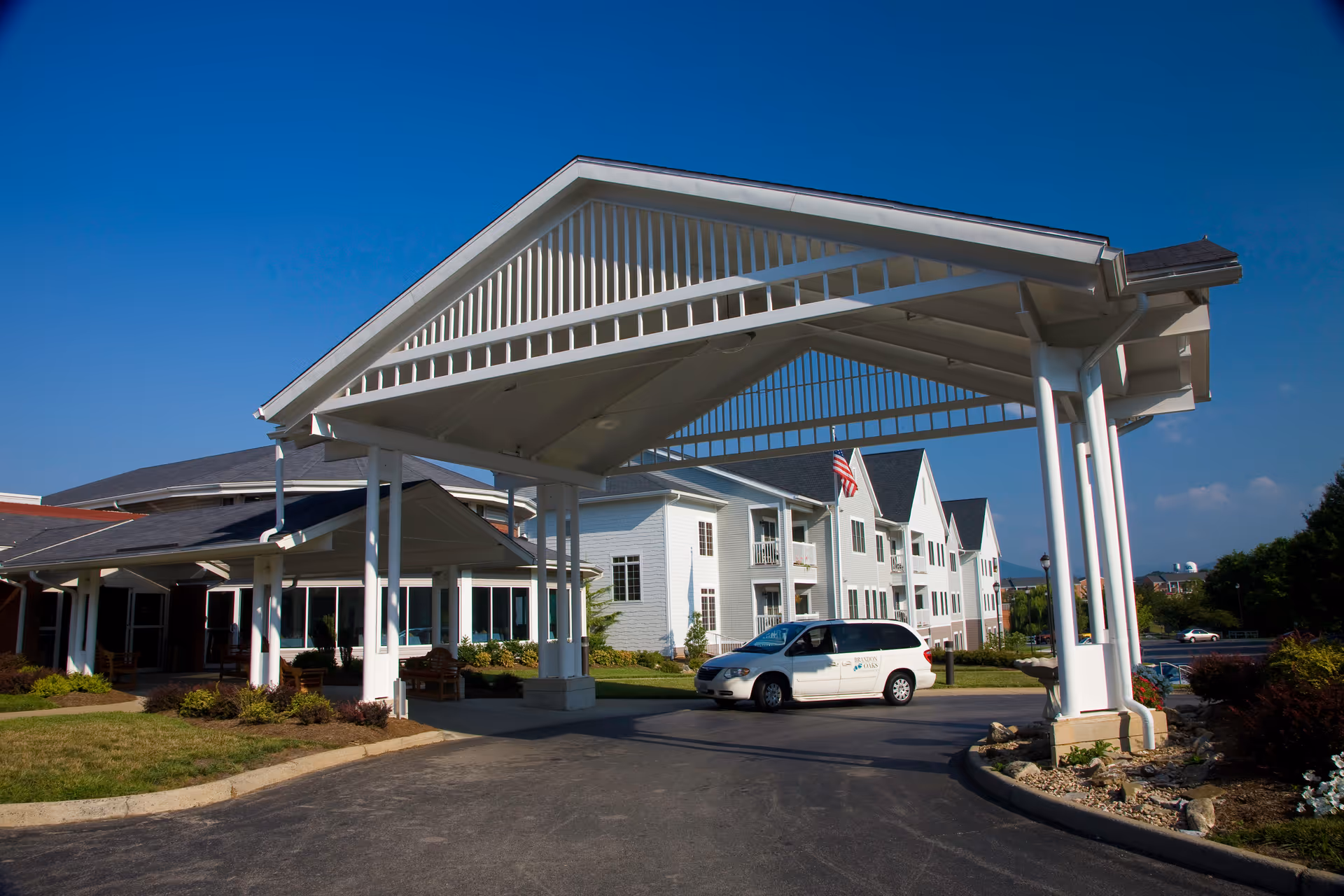 Entrance of Brandon Oaks Life Plan Community featuring a large white covered driveway with a white van parked underneath. The building is white with multiple windows and balconies, and an American flag is visible. The sky is clear and blue.