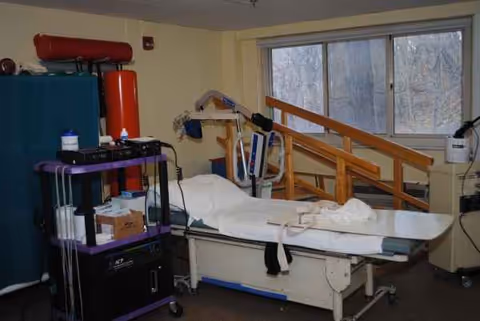 A hospital-style patient bed in a rehab room with equipment and a window.