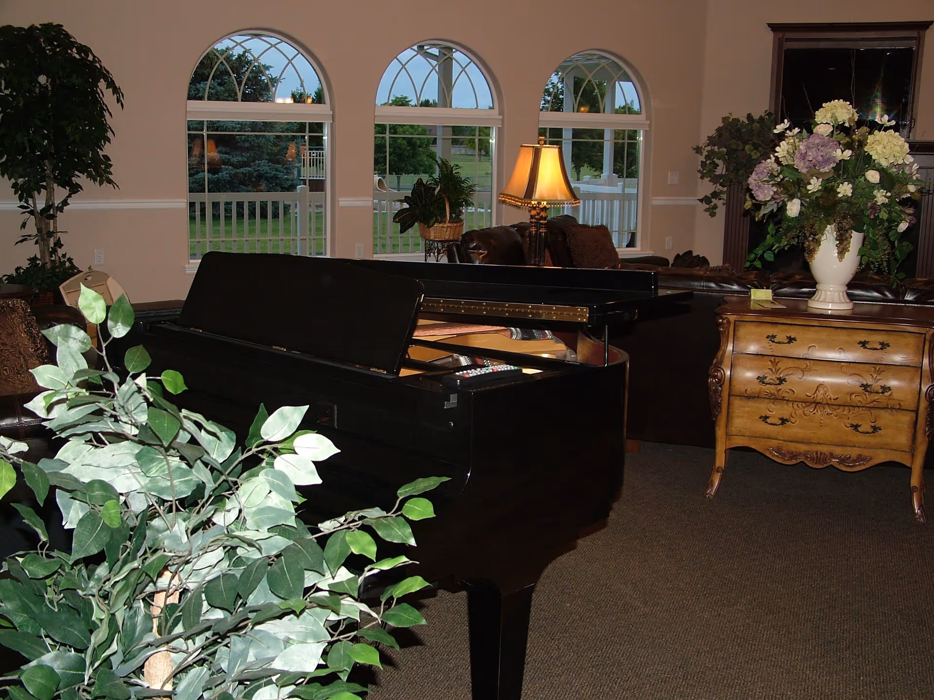 A furnished living room common area with a black grand piano in the foreground, potted plants, a decorative chest with a vase of flowers, and arched windows showing the outside.