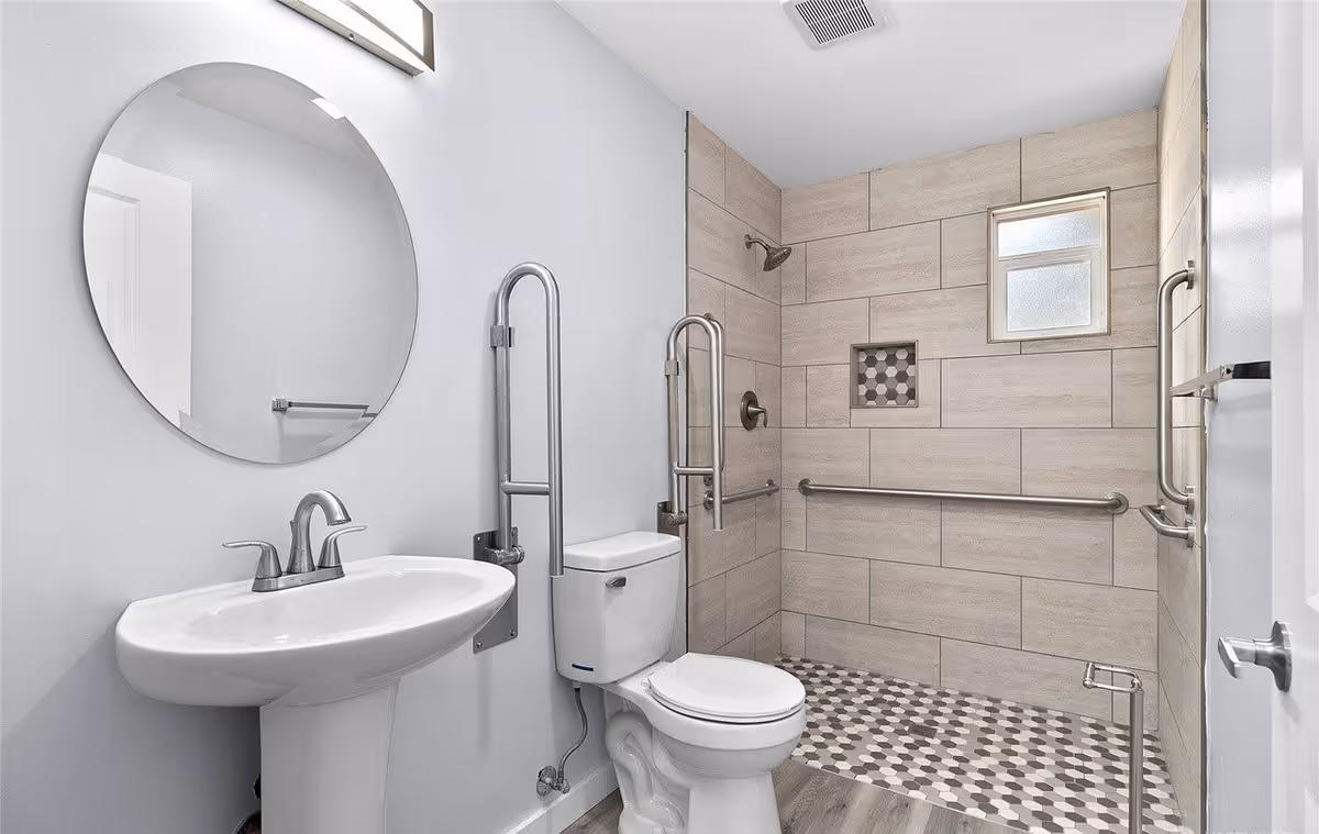 A modern accessible bathroom with a white pedestal sink, a round mirror above it, a toilet with grab bars on both sides, and a walk-in shower area with beige tiles and a small frosted window. The shower floor has a hexagonal tile pattern and there are multiple grab bars for support.