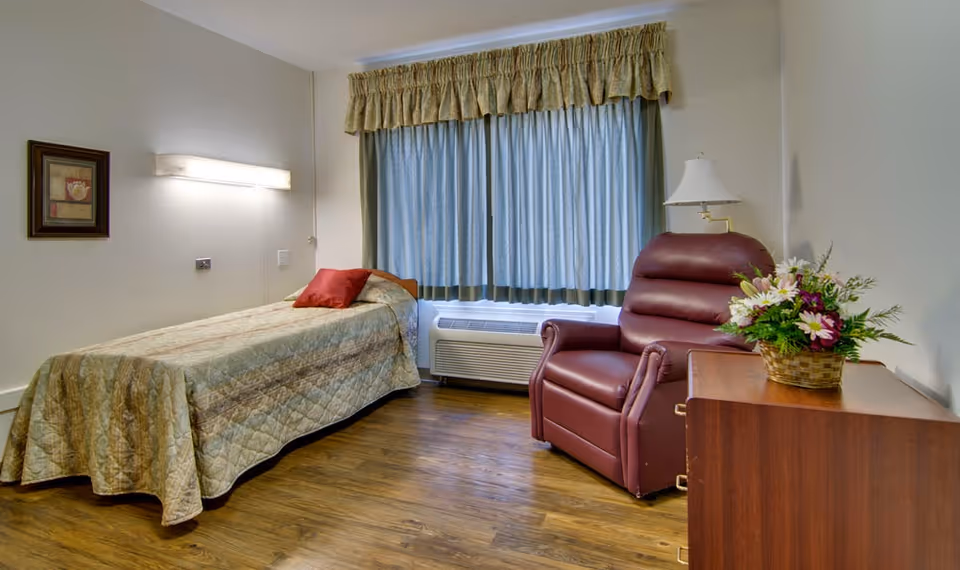 A simple bedroom with a single bed covered in a beige quilt and a red pillow. Next to the bed is a maroon recliner chair, a wooden dresser with a basket of flowers on top, and a floor lamp. A window with blue curtains and a beige valance is behind the chair. The floor is wooden, and there is a framed picture on the wall above the bed.