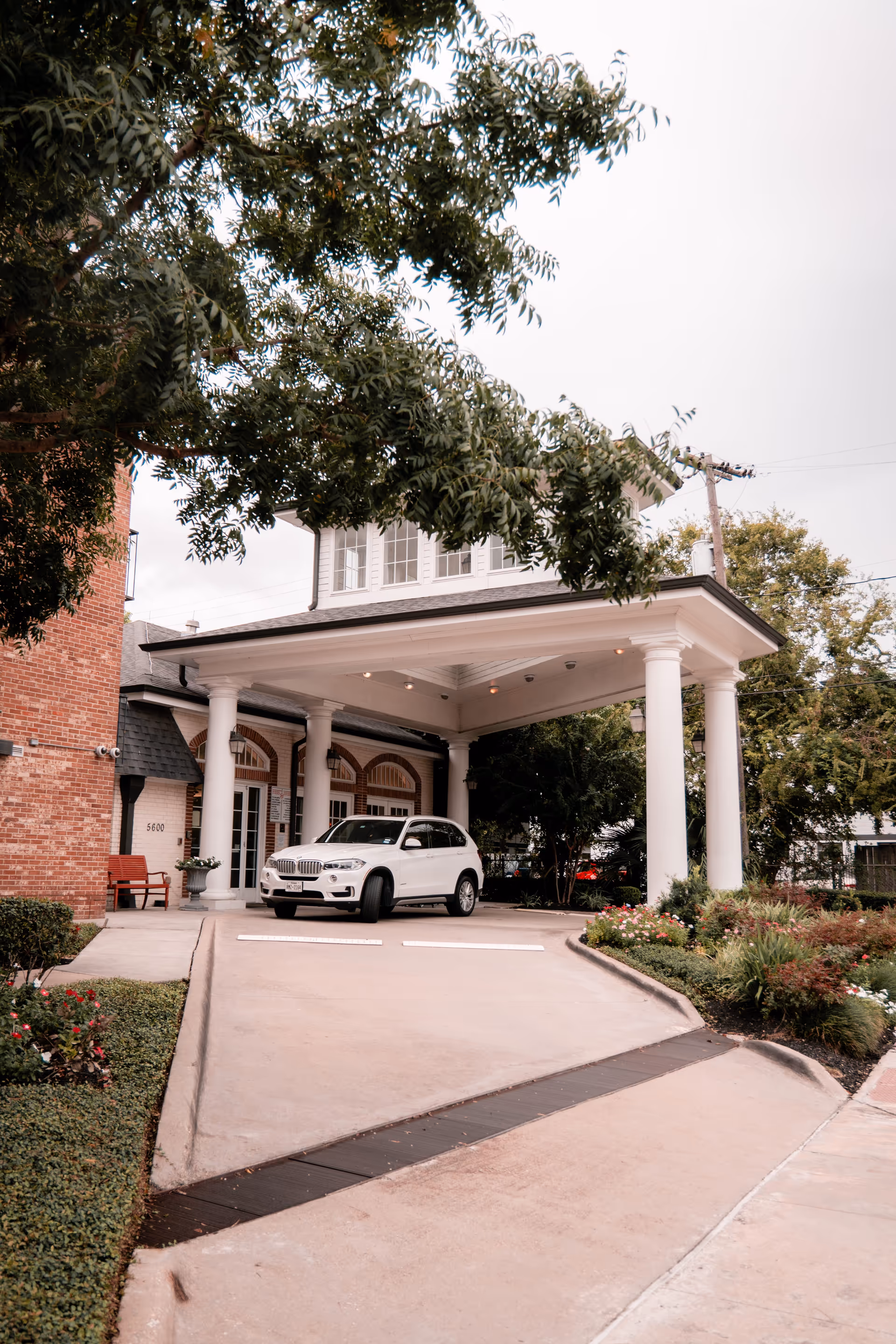 Front entrance of a brick building with a covered porte-cochere supported by white columns and a white SUV parked beneath, surrounded by trees and landscaping.