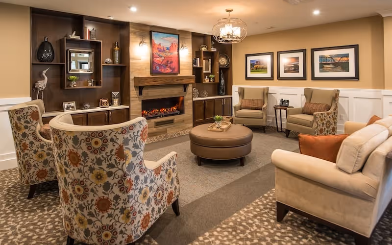 A cozy living room area with a modern electric fireplace set into a wooden accent wall. The room features a mix of patterned and solid upholstered armchairs and a beige sofa arranged around a round leather ottoman. The walls are decorated with framed landscape artwork and built-in shelves holding decorative items. The lighting includes wall sconces and a ceiling chandelier, creating a warm and inviting atmosphere.