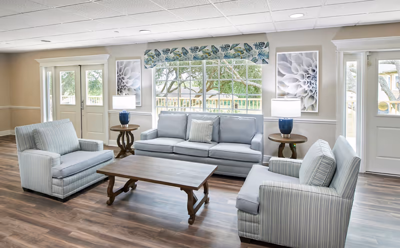 Bright communal living room with a light-blue sofa and two striped armchairs arranged around a wooden coffee table beneath a large window.