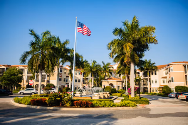 Exterior view of a senior living facility with a circular driveway featuring a central fountain surrounded by palm trees and landscaped greenery. An American flag flies on a flagpole in the middle of the fountain area. The building has a beige facade with red-tiled roofs and multiple windows. Several cars are parked along the driveway under a clear blue sky.