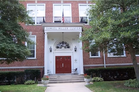 Front view of a brick building entrance with white columns, red double doors, and steps flanked by greenery.