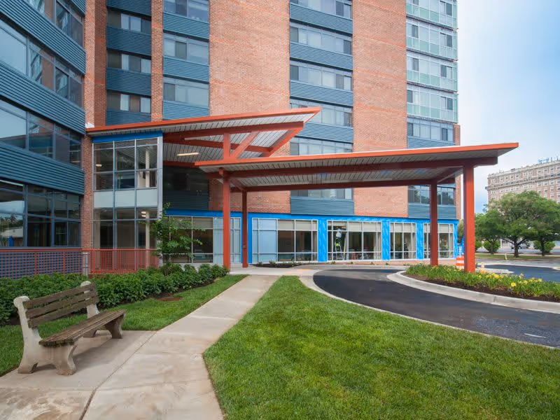 Entrance area of a multi-story brick and glass building with a covered drop-off zone supported by red beams. There is a concrete pathway leading to the entrance, bordered by green grass and shrubs. A wooden bench is placed along the pathway, and a tree is visible in the background near the parking area.