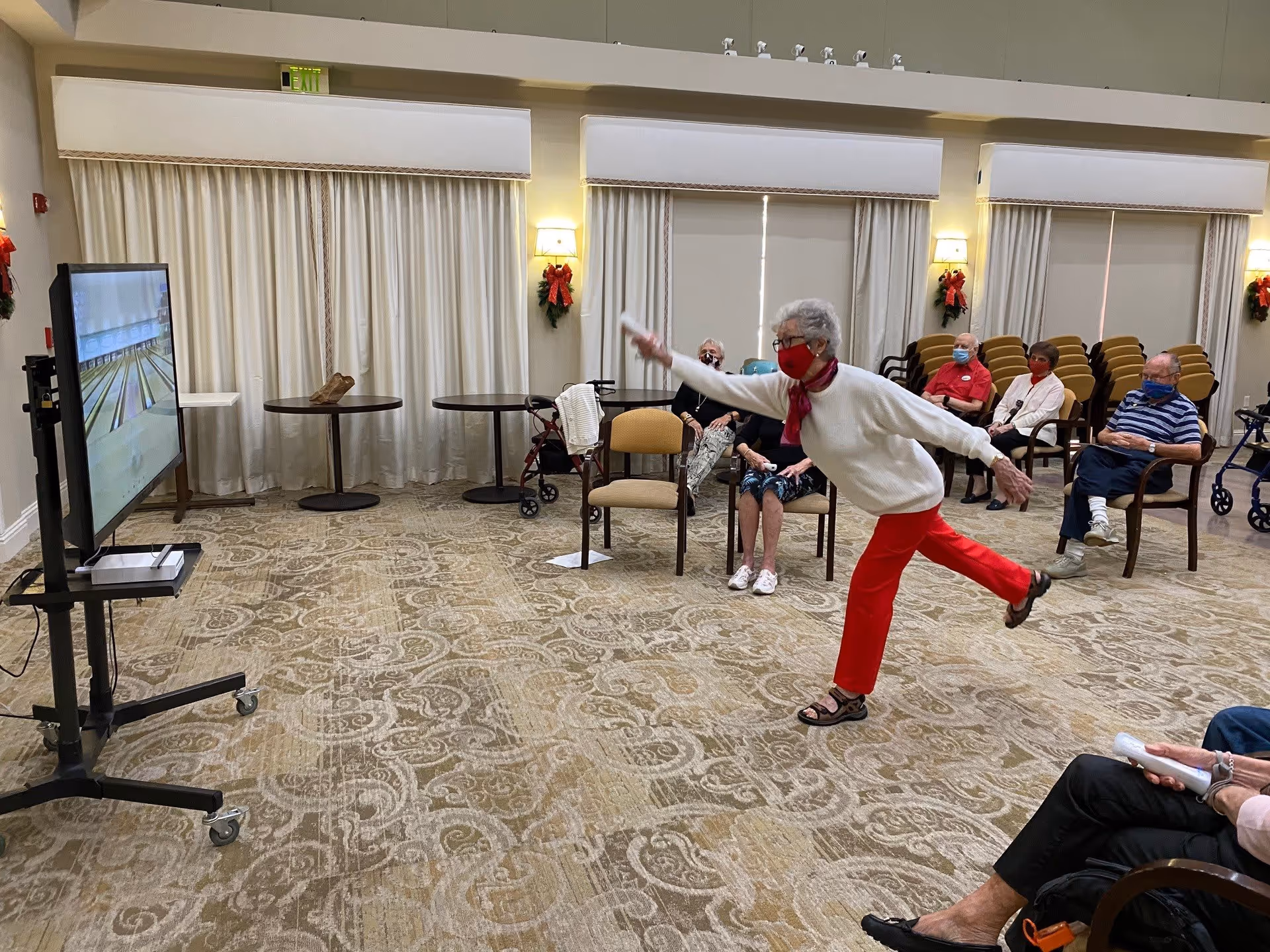 An elderly woman wearing a white sweater, red pants, and a red face mask is playing a virtual bowling game on a large screen in a spacious room. Several other elderly people are seated in chairs around the room, watching her play. The room has patterned carpet, cream-colored walls, and large windows covered with curtains. Some holiday wreath decorations are visible on the walls.