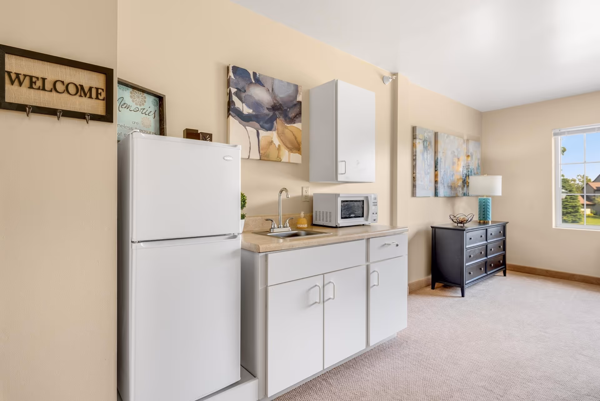 A small kitchenette area with a white refrigerator, white cabinets, a countertop with a sink, and a microwave. The walls are beige with framed artwork and a decorative 'WELCOME' sign. In the background, there is a black dresser with a lamp and additional artwork on the wall, next to a window showing a view of greenery outside.