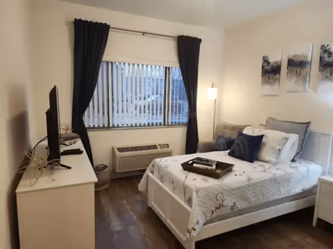A cozy bedroom with a white bed frame and floral-patterned bedding. The bed has multiple pillows and a wooden tray on top. There is a white dresser with a flat-screen TV on it to the left, a window with vertical blinds and dark curtains in the center, and a white nightstand with a lamp on the right. The floor is wooden, and three abstract paintings hang on the wall above the bed.