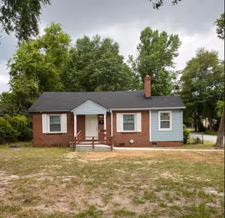 Single-story brick house with a small front porch, front lawn, and trees behind it.