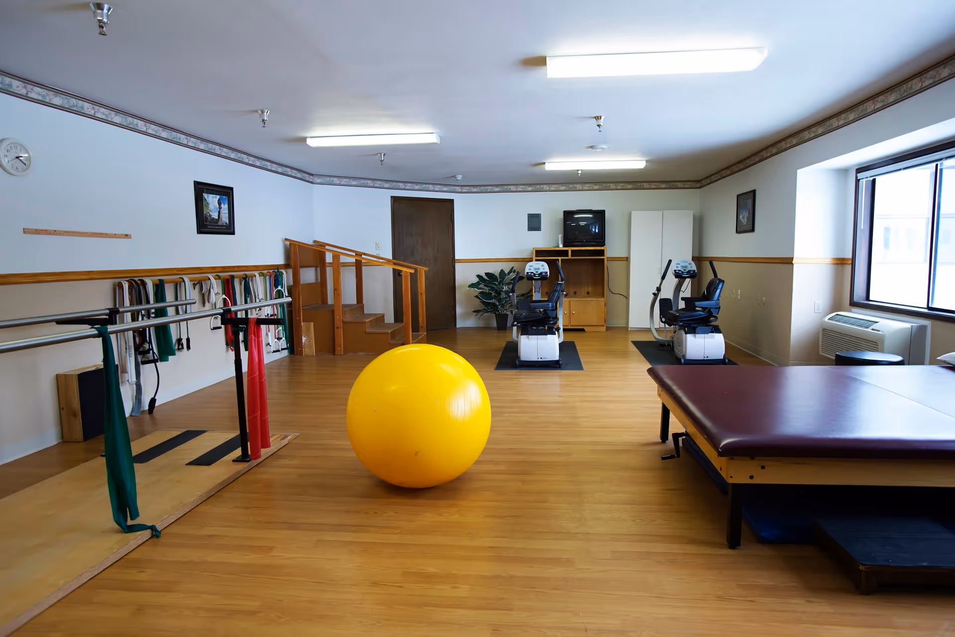 A physical therapy or exercise room with wooden flooring, a large yellow exercise ball in the center, two stationary exercise bikes near the back wall, a padded therapy table on the right, parallel bars with resistance bands on the left, and a small set of wooden stairs. The room has white walls with a wooden trim and a window on the right side letting in natural light.