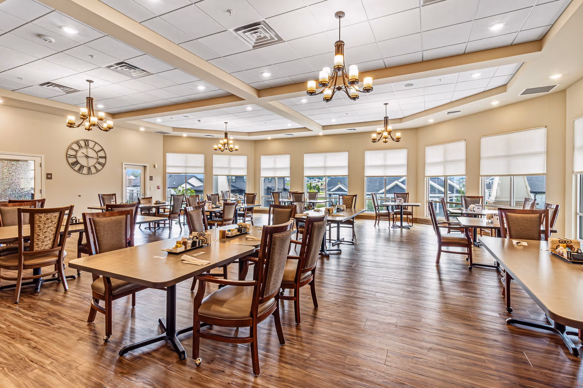 A spacious, well-lit dining room with multiple wooden tables and chairs arranged neatly. Large windows with white blinds allow natural light to fill the room. Several chandeliers hang from the ceiling, and a large wall clock is visible on one wall. The floor is covered with wood-style flooring.