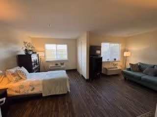 A spacious room with a bed on the left side, a dark wooden dresser, and a window with blinds. On the right side, there is a green sofa, a small table with a lamp, and another window with blinds. The floor is dark wood, and the walls are light-colored.