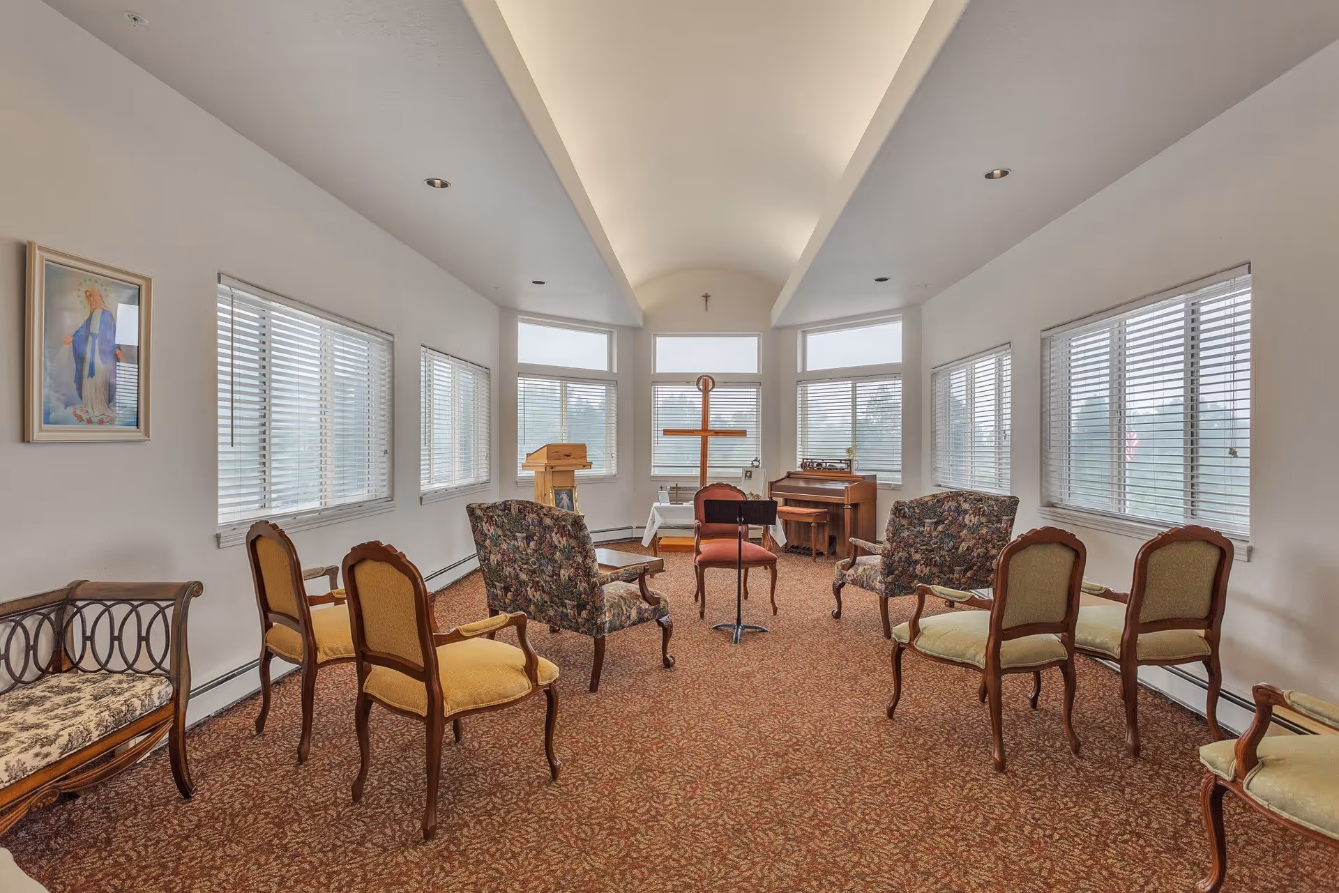 A small chapel or prayer room with multiple chairs arranged in a semi-circle facing a wooden cross on a stand. The room has large windows with blinds, a piano, a lectern, and a carpeted floor with a patterned design. A framed religious picture hangs on the left wall.