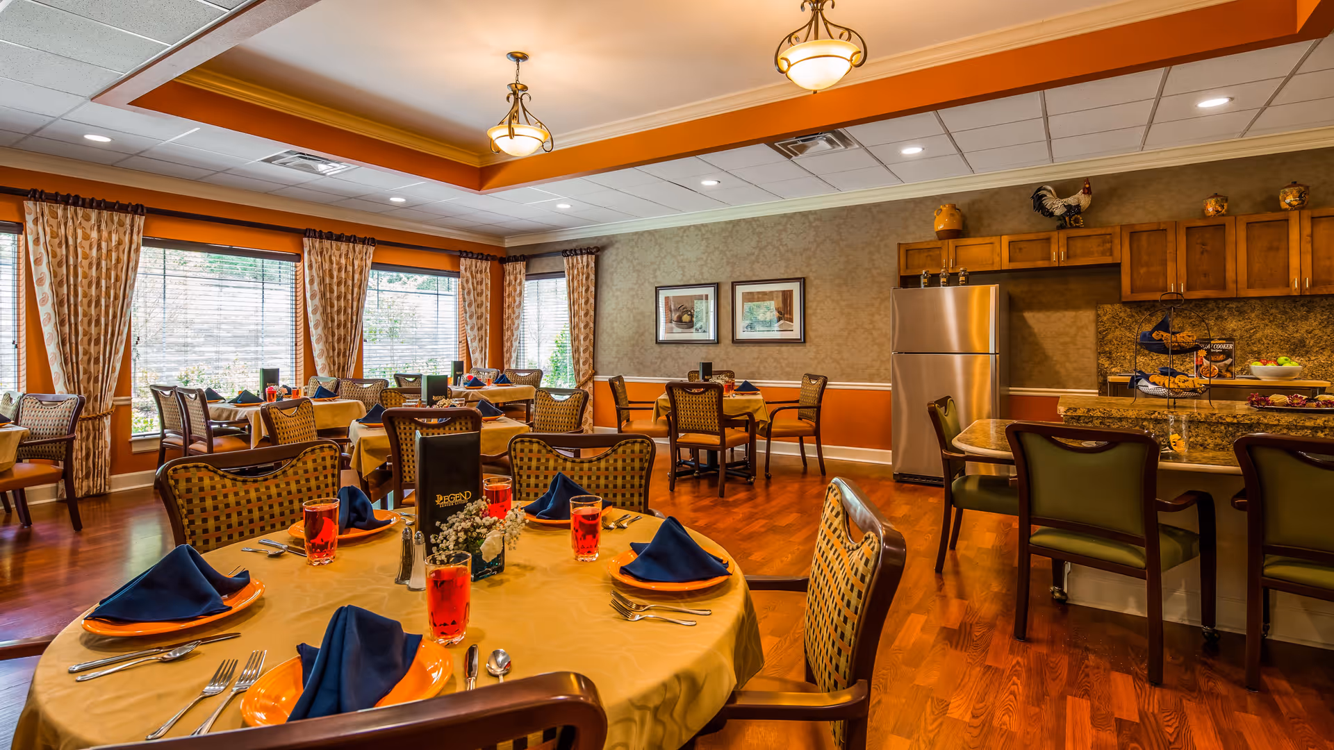 A warmly lit dining room with several tables set for a meal. Each table has yellow tablecloths, orange plates, blue napkins, and glasses filled with a red beverage. The room features large windows with patterned curtains, wooden chairs, and a kitchen area with a stainless steel refrigerator and granite countertops. The walls are decorated with framed pictures and ornamental items.