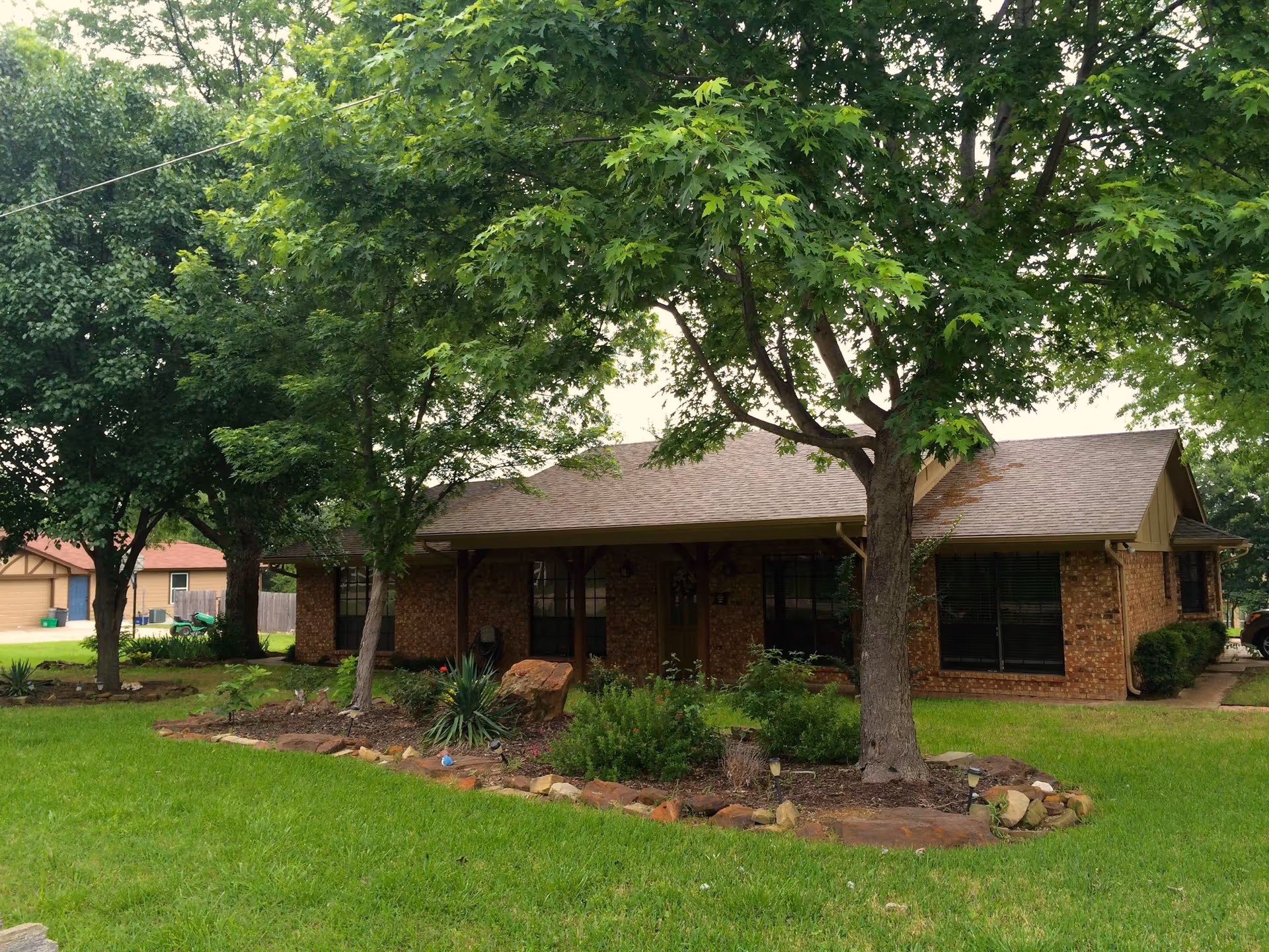 Single-story brick house with a front porch, large trees, and a landscaped lawn.
