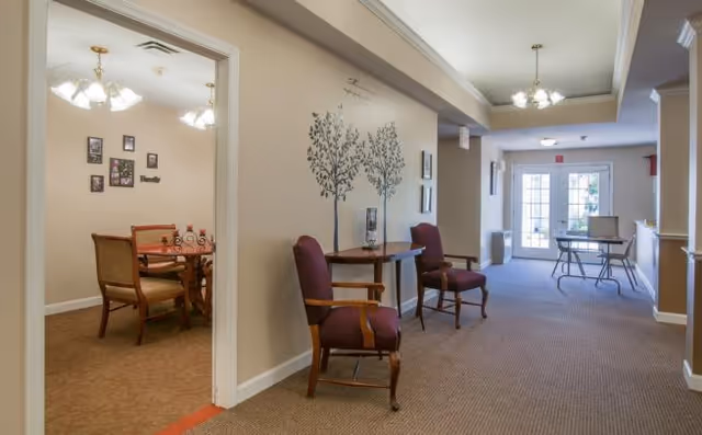 Interior hallway of a senior living facility with two wooden chairs with purple cushions placed against the wall. The wall features decorative tree decals and framed pictures. To the left, there is an open doorway leading to a dining room with a wooden dining table and chairs. At the end of the hallway, there are glass doors letting in natural light and a small table with chairs near the doors.