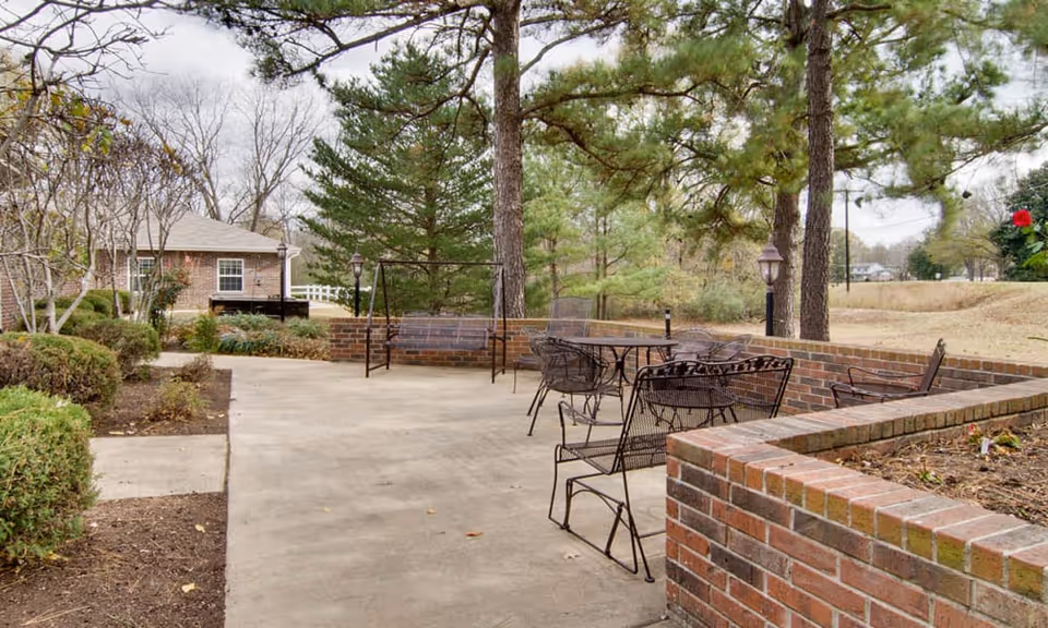 Outdoor patio with metal tables and chairs surrounded by low brick planters and pine trees.