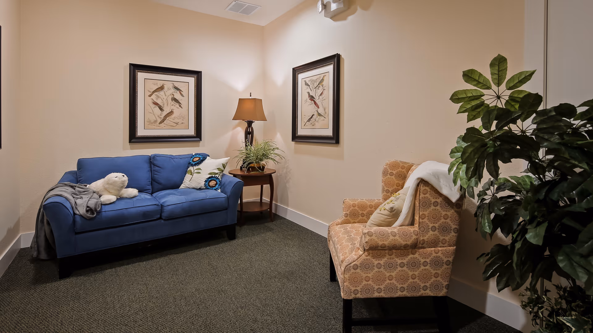 A cozy sitting area with a blue couch adorned with pillows and a stuffed white seal toy, an orange patterned armchair with a pillow and a white throw, a wooden side table with a lamp and a potted plant, two framed bird illustrations on the walls, and a large leafy green plant in the corner.
