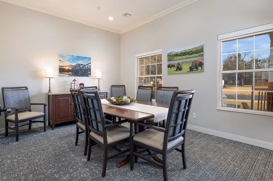 A dining room with a rectangular wooden table surrounded by six cushioned chairs. The room has two large windows letting in natural light, two framed landscape pictures on the walls, a small cabinet with two lamps and decorative items, and a carpeted floor.