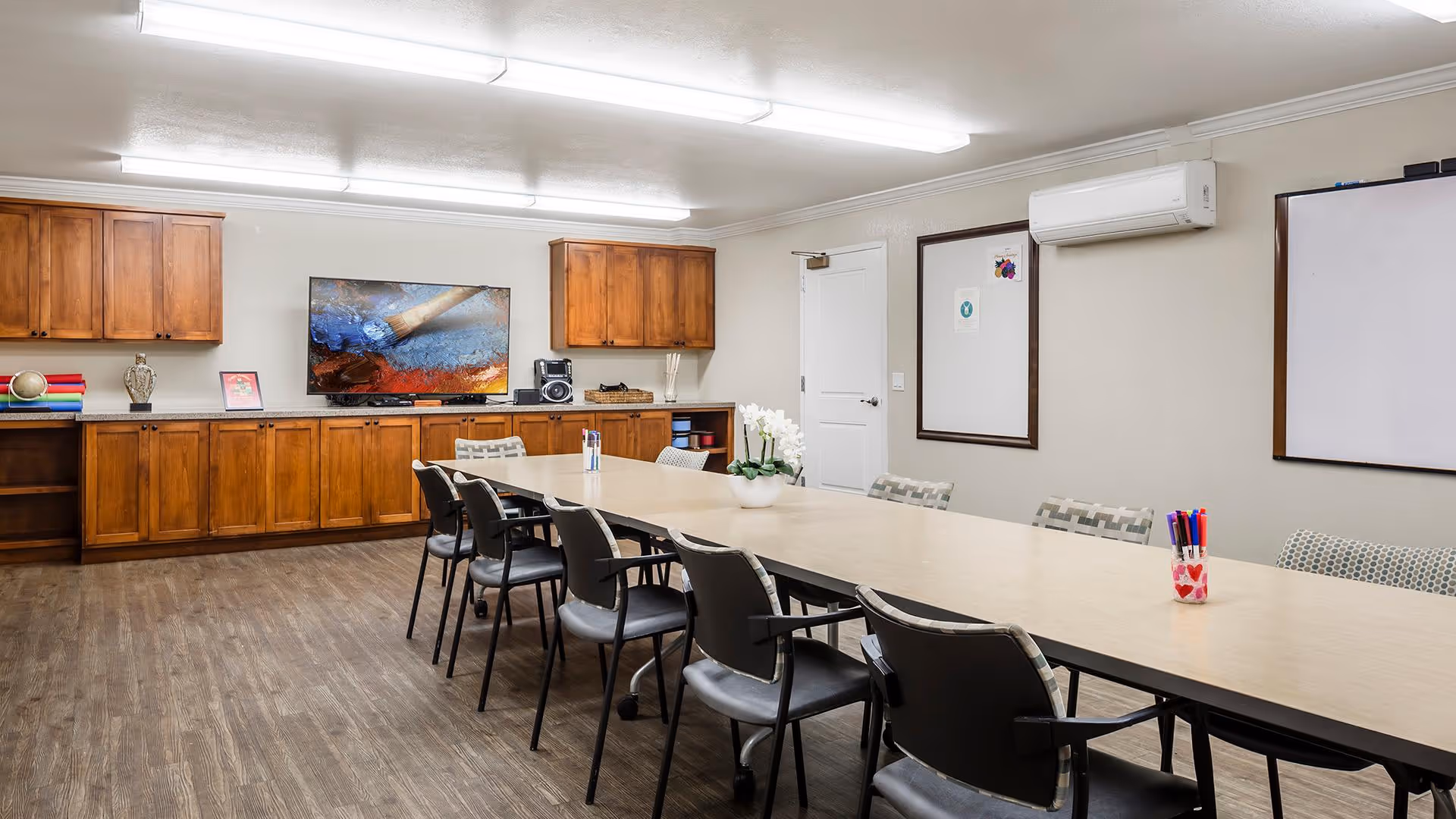 A well-lit conference or meeting room with a long rectangular table surrounded by chairs. The room has wooden cabinets along one wall, a flat-screen TV mounted above the cabinets, and two whiteboards on the opposite wall. There is a white door and an air conditioning unit on the wall. The table has a small plant and a container with colorful markers.