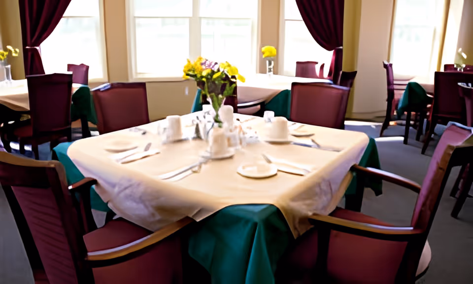 A dining room with multiple tables covered in green and beige tablecloths, each set with white cups, plates, utensils, and a centerpiece of yellow flowers. The room has large windows with white blinds and dark red curtains, and several wooden chairs with red cushions are arranged around the tables.