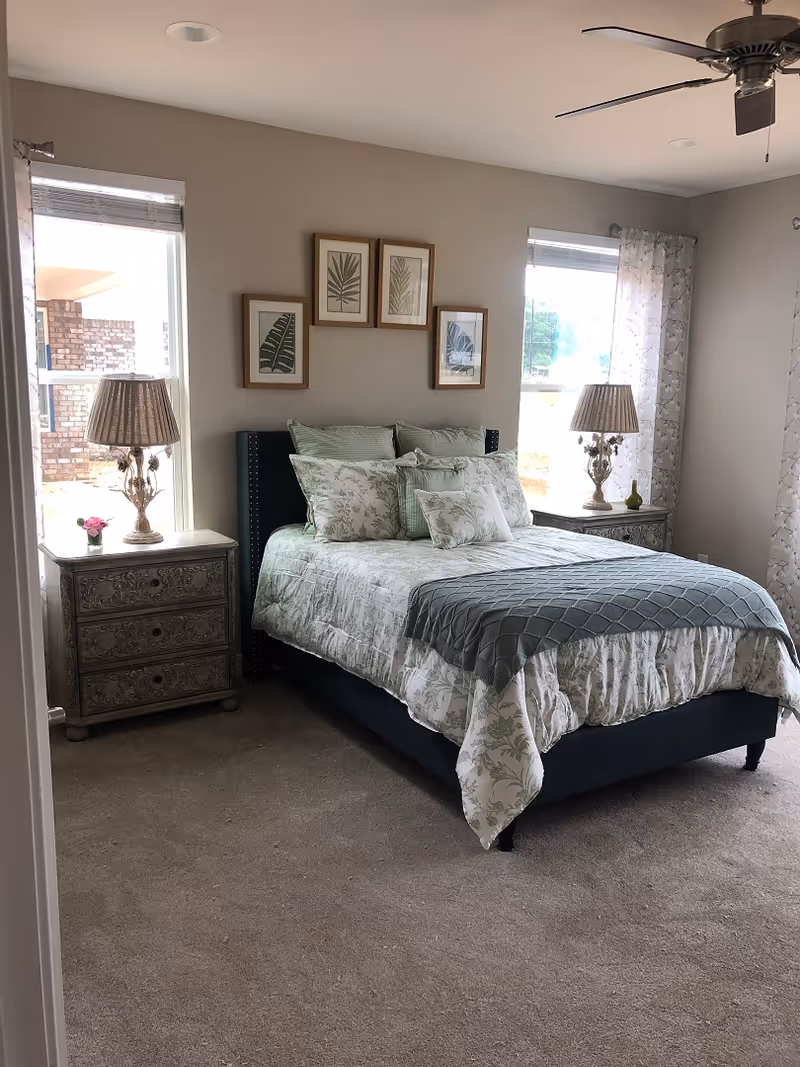A neatly made bed with floral and green patterned bedding and multiple pillows in a bedroom. There are two windows on either side of the bed with sheer curtains and blinds. Each side of the bed has a decorative nightstand with a lamp. Above the bed, there are five framed botanical prints arranged in a pattern on the wall. The room has beige carpet and a ceiling fan with light.