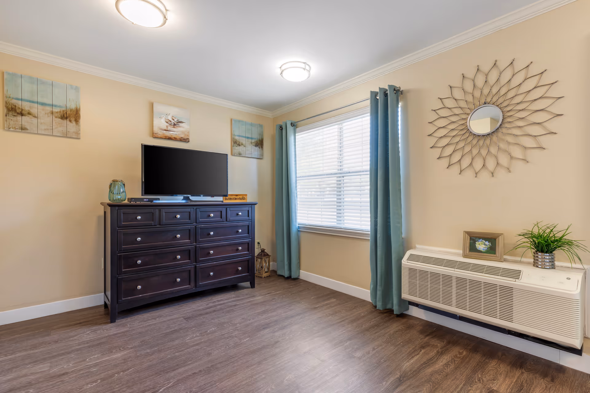 Bright sitting area with a dark dresser and TV opposite a window with teal curtains and a decorative sunburst mirror above a wall-mounted air unit.