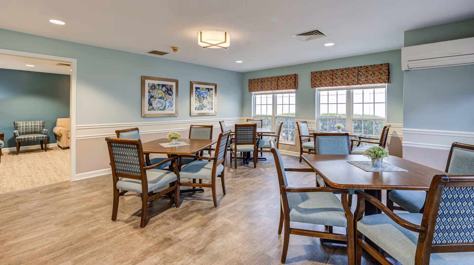 A bright dining room in a senior living facility with several wooden tables and cushioned chairs arranged neatly. The walls are painted light blue with white trim, and two framed floral paintings hang on one wall. Large windows with patterned valances let in natural light, and small flower arrangements decorate each table. An adjacent room with additional seating is visible through an open doorway.