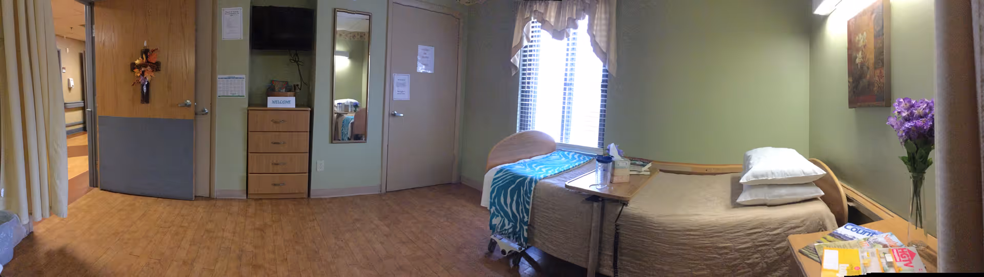 A senior living facility bedroom with a single bed covered in beige bedding and a blue patterned blanket at the foot. There is a small wooden dresser with a welcome sign, a wall-mounted TV, a mirror, and a closed door. A window with blinds and a valance lets in natural light. A small table is positioned over the bed, and a vase with purple flowers is on a side table next to the bed. The floor is wood, and the walls are painted light green.